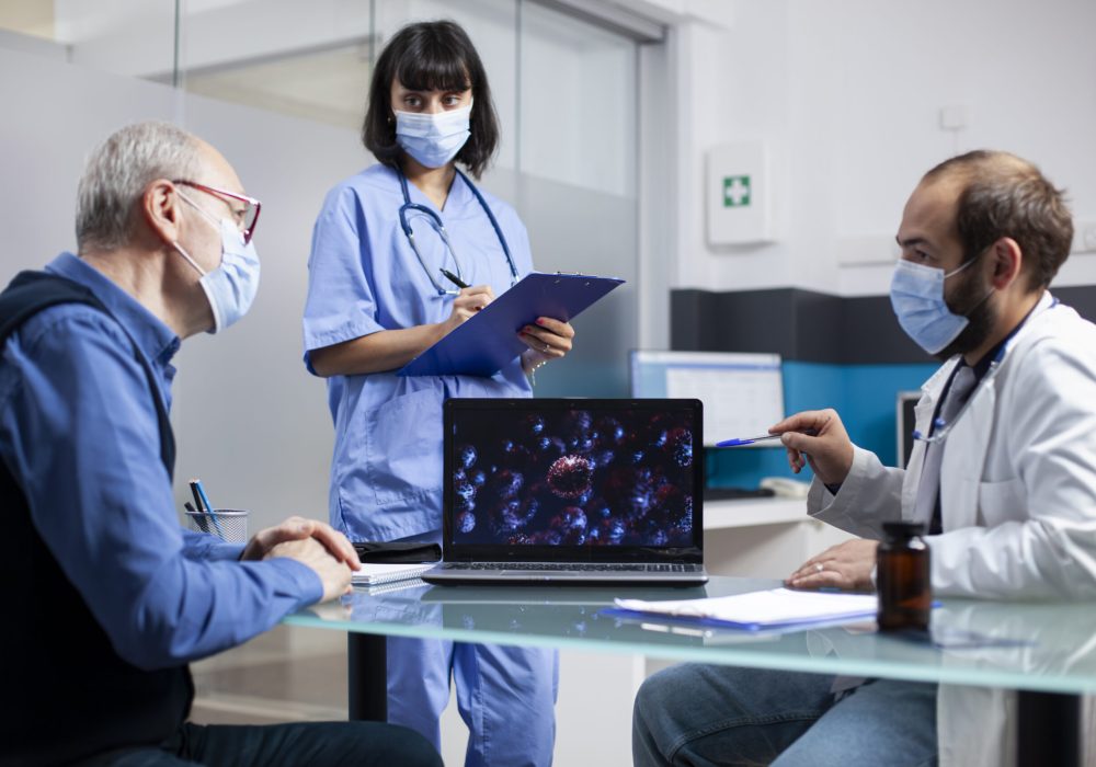 Doctor and old patient analyze digital display of microscopic germs as female nurse writes notes on clipboard. Healthcare workers discuss disease prevention and treatment with senior man in clinic.